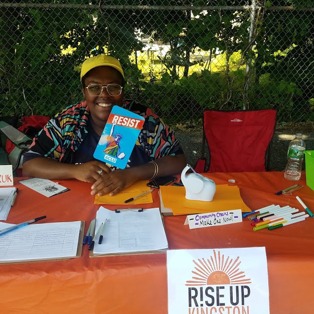 Image of a person sitting and smiling behind an orange table with a sign that says Rise Up Kingston in the front