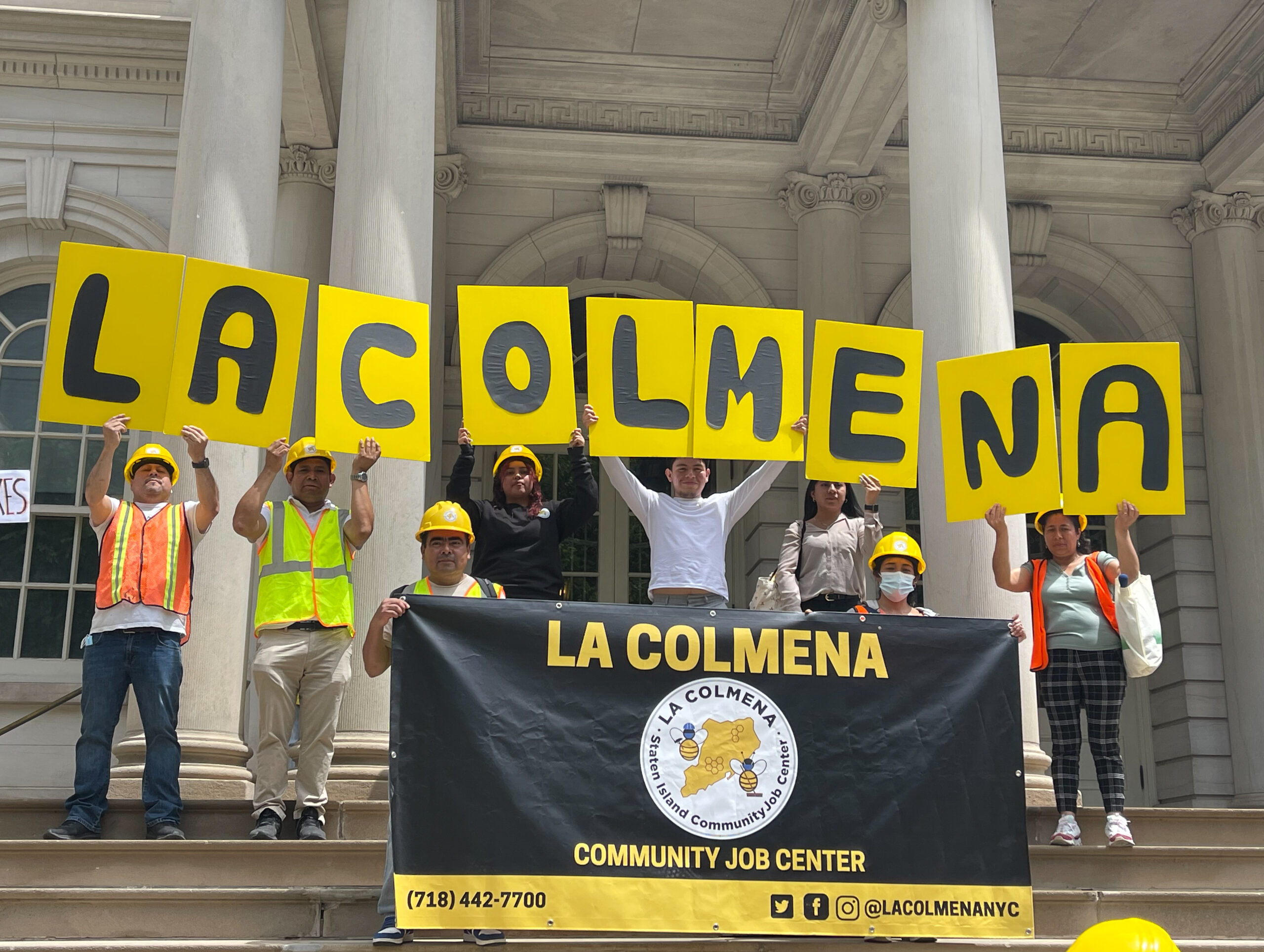 Members of La Colmena stand outside the steps of a government building holding signs that read "La Colmena"