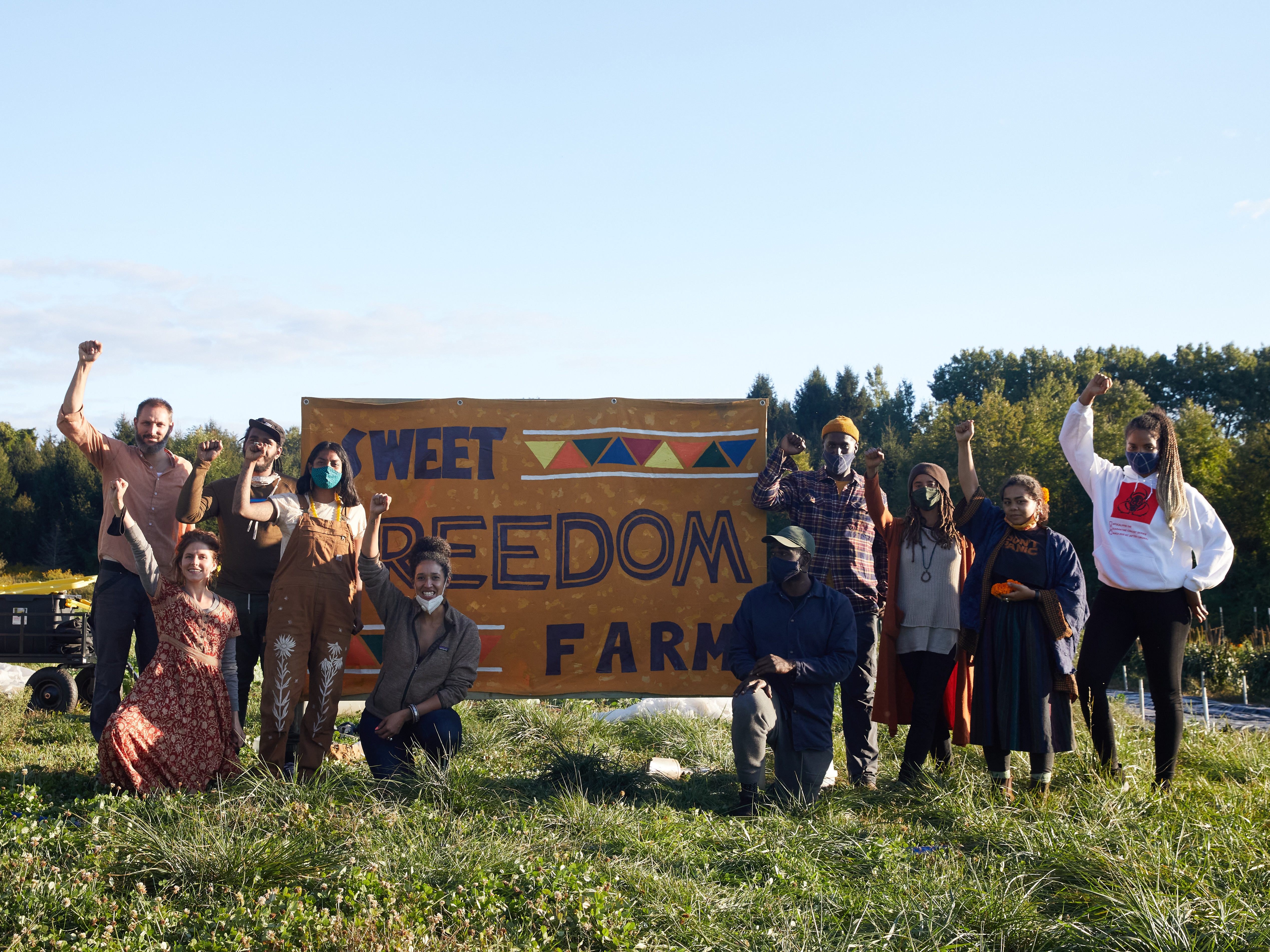 A group of people stand in front of a sign reading Sweet Freedom Farm, some with their fists to the sky